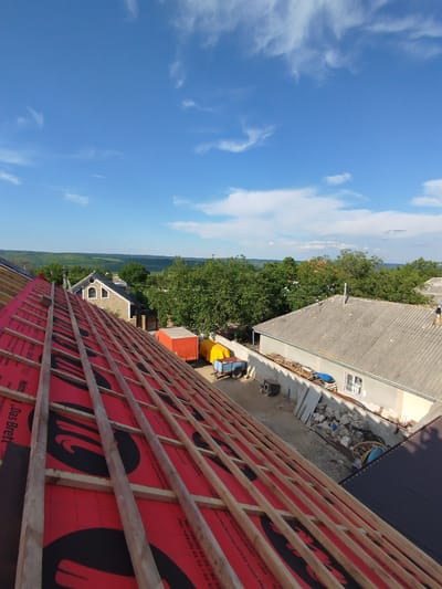 This is a medium-shot, eye-level, vertical-orientation photograph taken from a rooftop, looking out over a rural residential area. The foreground is dominated by a rooftop under construction, featuring exposed wooden rafters laid over bright red roofing underlayment. The underlayment has a repeating black logo or pattern and some German text, likely product information, with phrases like "Das Brett" and "Markenqualität."

Beyond the immediate rooftop, the scene transitions to a village with several houses. One house in the middle ground is built of stone with a dark roof and decorative arched windows. Further to the right, a lighter-colored house with a gray, tiled roof is visible. Several trees, lush and green, dot the landscape, suggesting it is summer.

In the yard between the houses, various construction-related objects are visible: an orange shipping container, a blue trailer with a yellow tarp, piles of building materials including wood and stones, and some coiled hoses or cables. The sky above is a vibrant blue, punctuated by scattered white, wispy clouds, indicating it is daytime, likely late morning or early afternoon. The overall impression is one of ongoing renovation or construction work in a peaceful, rural setting in Pănășești, Moldova.
