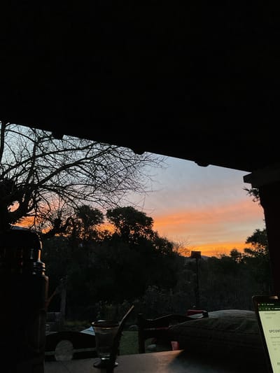 The image captures a serene outdoor scene at dusk, likely in El Rodeo, Argentina. In the foreground, a dark thermos and a glass with a straw are placed on a wooden surface. To the right, a laptop displays a webpage with text in Spanish, including "OPCIONES". The background reveals a silhouette of trees and vegetation against a sky painted with the warm hues of sunset, transitioning from orange to soft blue. The overall atmosphere is tranquil and suggestive of a quiet evening at home, perhaps on a patio or porch, with the natural beauty of the El Rodeo landscape providing a picturesque backdrop.