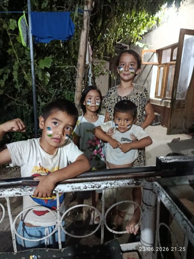 Four children, appearing to be of Central Asian descent, are posed for a photograph. Three of them, two boys and one girl, have face paint resembling the flag of Uzbekistan. The youngest boy in the foreground is flexing his bicep. He is wearing a white t-shirt with a graphic print and blue shorts. To his right stands a younger girl, also with face paint, wearing a white t-shirt with a floral pattern. Behind them, a slightly older girl, the tallest of the group, wears a patterned dress and also has face paint. A partially visible adult, likely an elder, is standing behind the younger girl.
The scene is outdoors at night, suggested by the artificial lighting and the dimness of the surroundings. There are trees and foliage in the background, and what appears to be a rustic outdoor structure with wooden windows and doors. A white metal railing is in the foreground, which the children are leaning on or standing behind. The overall atmosphere seems informal and celebratory, possibly related to a national event or holiday given the face paint.
The visible text on the image is a date stamp: "23.06.2026 21:25".