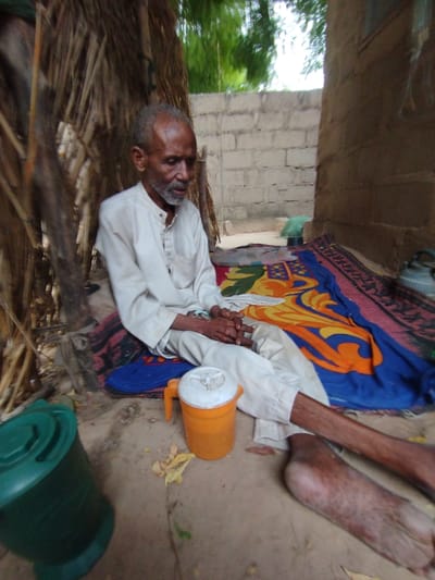 The image shows an elderly man sitting on the ground in Bajoga, Nigeria, near a brick wall, a thatch roof, and a colorful blanket. He is wearing a white long-sleeved shirt and white pants. He is sitting with his legs crossed, his feet out in front of him. He is holding his hands in his lap. There is an orange pitcher with a white lid next to him.  There is also a green container on the ground to his left.  The environment is dusty and dry.  The background is blurred. The coordinates provided, 10.851 latitude and 11.446 longitude, correspond to a rural area within Bajoga, Nigeria.