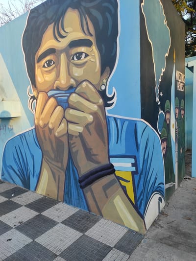 The image shows a mural in Remedios de Escalada, Argentina. It depicts a stylized portrait of Diego Maradona, a famous soccer player, wearing a blue and white jersey, holding it up to his face. Above Maradona's portrait is a map of Argentina with the words "Nuestros Héroes" ("Our Heroes") painted next to a depiction of Argentinian soldiers with an Argentinian flag. The mural is painted on a wall, and a tiled sidewalk with a black and white checkered pattern is visible in the foreground.