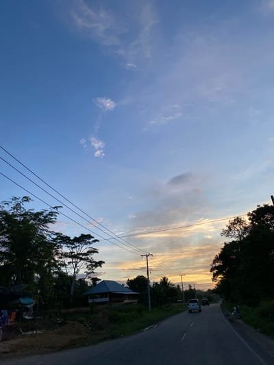 This image captures a road at sunset in Kutameuligoe, Indonesia. The sky is a gradient of blue, with streaks of orange and yellow clouds near the horizon, indicating the setting sun. Power lines stretch across the frame, supported by utility poles. To the left, lush green trees and vegetation line the road, with a simple house or structure partially visible amongst them. A small settlement with some buildings and objects, possibly merchandise, is also present on the left side. On the right side of the road, more trees and foliage form a dense border. Several vehicles are on the road: a silver SUV, a darker car behind it, and a parked motorcycle on the right shoulder. The road surface is asphalt and appears to be in good condition, with visible lane markings. The overall atmosphere suggests a rural or semi-rural setting experiencing the transition from day to night.
