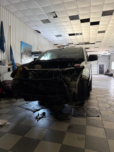 The image shows the interior of what appears to be a workshop or garage in Parma, Russia. The primary subject is a white van that is heavily damaged, with its front end dismantled, exposing the engine and internal components. The van is positioned in the center of the frame, occupying a significant portion of the space.

The setting is characterized by a tiled floor with a checkerboard pattern of light and dark gray tiles. The walls are lined with white panels, some of which show signs of damage or wear, with sections missing or torn. The ceiling is also made of white panels, with some grid-like structures visible.

Several objects are scattered around the workshop. To the left of the van, leaning against the wall, are several long wooden poles, possibly part of a tool or equipment. There are also some blue objects, possibly flags or banners, with some visible text on them, though it is difficult to decipher. On the wall behind the van, a framed picture or poster is visible, depicting a scene with a motorcycle.

In the background, a doorway is visible, suggesting an exit or another part of the facility. There are also some pieces of equipment or debris on the floor near the van. The lighting in the image appears to be artificial, likely from overhead lights, and casts shadows on the floor. The overall impression is one of a functional but somewhat cluttered workspace where repairs or dismantling are taking place. There are no people visible in the image. The time of day is not evident, and there is no indication of weather conditions.