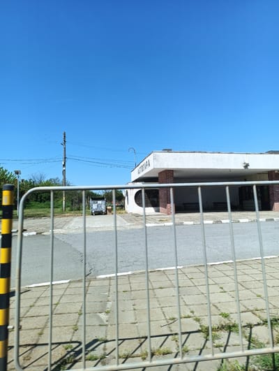 The image shows the Avtogara (bus station) of Zimnitsa, Bulgaria, viewed through a metal fence. The building is a single-story, white structure with brick support columns. The word "АВТОГАРА" is written in large letters on the building's facade. The area in front of the station is paved with asphalt and square concrete tiles. A dumpster is visible near the building. A utility pole with wires is on the left. The sky is clear and blue.
