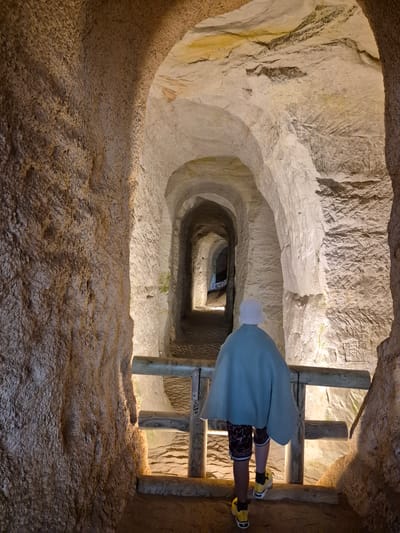A person wearing a light blue cloak and a white hat walks through a man-made cavern. The cavern walls are rough and textured, with rough-hewn arches creating a tunnel-like passage that recedes into the distance. Wooden railings line the path. The scene appears to be inside the Piusa Sand Caves in Piusa, Estonia, a network of underground tunnels excavated for glass sand.