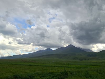The image captures a panoramic view of a pastoral landscape near Toguz-Bulak, Kyrgyzstan. The scene is dominated by rolling green hills that transition into a majestic mountain range in the background. Several of the peaks are snow-capped, indicating a high altitude. The sky is filled with dramatic, heavy clouds, with patches of blue sky peeking through, suggesting dynamic weather conditions. In the foreground, there are fields of green grass and cultivated land. A few scattered trees are visible in the mid-ground, as well as a lone vehicle. The overall impression is one of vast, natural beauty and a sense of rugged wilderness.