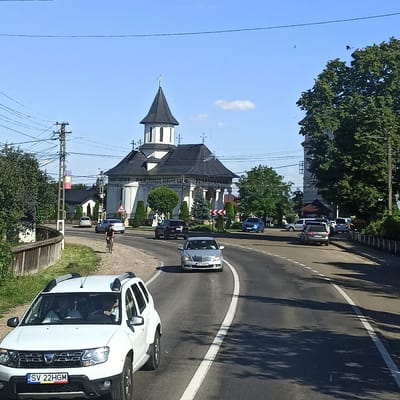 The image captures a sunlit street scene in Milișăuți, Romania, featuring a prominent white church with a dark, multi-tiered roof and a steeple topped with a cross. The church is flanked by trees and smaller buildings. A two-lane asphalt road curves through the scene, with a white line delineating the lanes. Several vehicles are visible: a white Dacia Duster SUV in the foreground with Romanian license plates "SV 22HGM," a silver Mercedes-Benz sedan behind it, a dark SUV, and a blue car further down the road. A cyclist, a person wearing dark clothing, is riding a bicycle on the right side of the road. Power lines and poles are a significant feature of the environment, particularly on the left side of the image. A wooden fence runs along the left edge of the road. The sky is clear blue with a few scattered clouds.