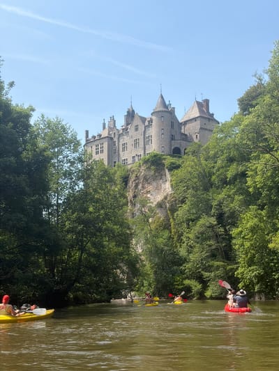 The image shows a view of the Château de Walzin in Dinant, Belgium, from a river perspective. The medieval castle is perched on a rocky cliff overlooking the Lesse River. The river contains several kayaks with people paddling along. The landscape is heavily wooded. The sky is blue with some contrails.
