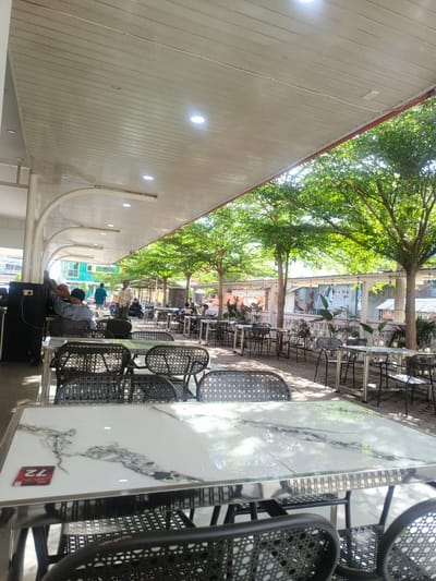 The image depicts an outdoor or semi-outdoor dining area, likely a cafe or restaurant, located in the city of Pantegajah, Indonesia. The scene is illuminated by bright daylight, with sunlight filtering through trees and casting shadows.

In the immediate foreground, multiple empty tables with glossy white marble-patterned tops and reflective chrome-finish frames are visible. Dark-colored chairs with woven, rattan-style backs and seats are positioned around these tables. A red square card marked "72" rests on the leftmost table. The table surfaces reflect portions of the sky and tree canopy.

The dining area extends under a flat, slatted white ceiling supported by white arched and straight columns. Recessed circular downlights and linear fluorescent light fixtures are integrated into the ceiling. Several individuals are present: two men are seated at a table on the left, one wearing a light blue shirt and a red cap, facing away from the camera. Further back, a man in a plaid shirt stands near a parked motorcycle. Other patrons are sparsely distributed at tables further into the covered area. A dark, rectangular speaker or equipment box is positioned on the far left, adjacent to a column.

Beyond the covered section, an open-air seating area is shaded by a line of mature trees with lush green foliage and slender trunks. More tables and chairs are arranged beneath these trees on a paved surface that displays dappled sunlight and shadows. A low, white-painted wall with decorative ventilation blocks separates this area from the background.

The background reveals an urban environment. Behind the wall, various structures are visible, including corrugated metal sheds and utilitarian buildings, some with laundry hanging. Further back, multi-story buildings with glass windows and varied architectural styles are present on the left, while more modest, possibly residential or commercial structures are on the right.