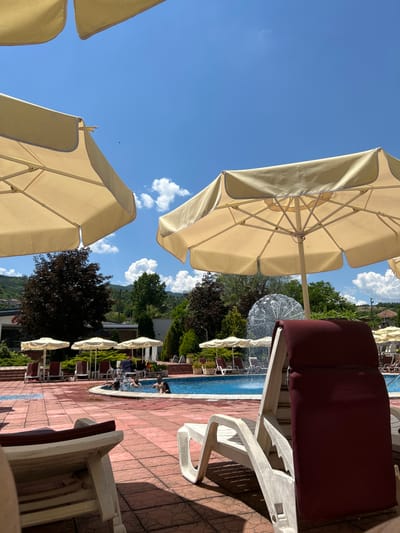 The image depicts a sunny day at an outdoor swimming pool located in Zverino, Bulgaria. In the foreground, two white plastic lounge chairs with burgundy cushions are positioned on a red tiled patio. The left chair is partially visible, while the right chair is fully in frame. Behind these chairs, a blue swimming pool is visible, with several people enjoying the water. To the left of the pool, a large, decorative fountain sprays water upwards. Several large, light-yellow umbrellas provide shade over the pool area and lounge chairs. In the background, lush green trees and rolling hills covered in vegetation form the natural landscape, with a few buildings visible amongst the trees. The sky is clear blue with scattered white clouds.