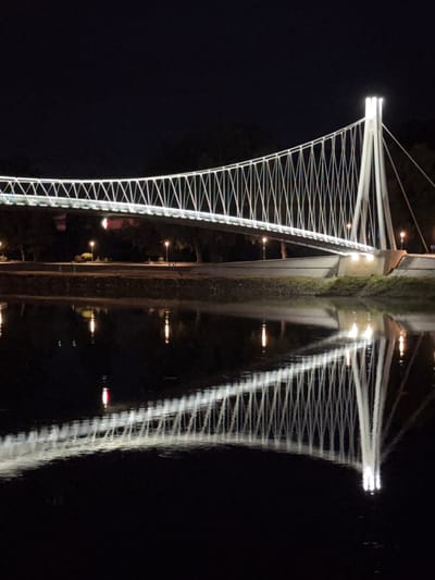 The image displays a modern pedestrian suspension bridge brilliantly illuminated at night, with its reflection clearly visible in the calm water below. The bridge, located in Osijek, Croatia, spans horizontally across the frame. It features two prominent, brightly lit pylons at its center, from which numerous thin, white cables extend outwards and downwards, forming a distinctive criss-cross pattern along the bridge's length. The main pathway of the bridge is also well-lit.
To the left of the bridge's main structure, a parallel pathway or bank is visible, adorned with several smaller, warm-toned lampposts and one notable red light, all of which are also reflected in the water. The surrounding environment is dark, highlighting the artificial illumination of the bridge and its immediate vicinity. The reflection of the bridge and its lights is almost symmetrical, though gentle ripples on the water's surface create subtle distortions, particularly elongating the reflected light sources and blurring the fine details of the cables. The scene conveys a tranquil nighttime atmosphere. There is no visible text in the image.