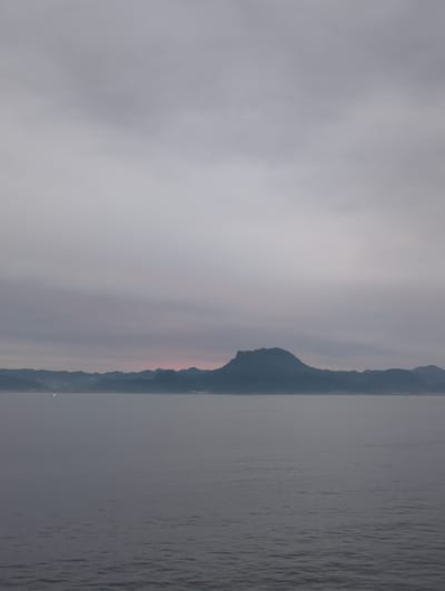A seascape at dusk shows a calm body of water in the foreground, with undulating waves under a gray sky. In the distance, a long, dark mountain range stretches across the horizon, with a prominent, rounded peak in the center. The coastline features a faint outline of buildings and lights. The atmosphere is subdued, with soft hues of pink and purple visible in the sky behind the mountains, suggesting either sunrise or sunset. The overall scene evokes a sense of tranquility and natural beauty.