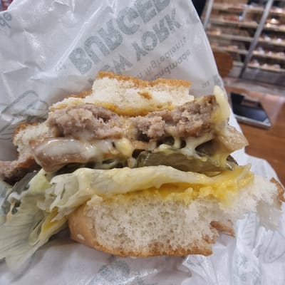 A close-up, eye-level shot captures a partially eaten burger, wrapped in white paper with embossed text that is partially visible. The burger, with its top bun removed, reveals layers of meat, melted cheese, lettuce, and pickles. The texture of the burger's ingredients, including the crumbled meat patty and gooey cheese, is evident. The background is softly blurred, showing shelves stocked with food items, suggesting a retail or food service environment. The lighting is bright, highlighting the food. The exact location within Wonju-si, South Korea, is not discernible from the image itself.