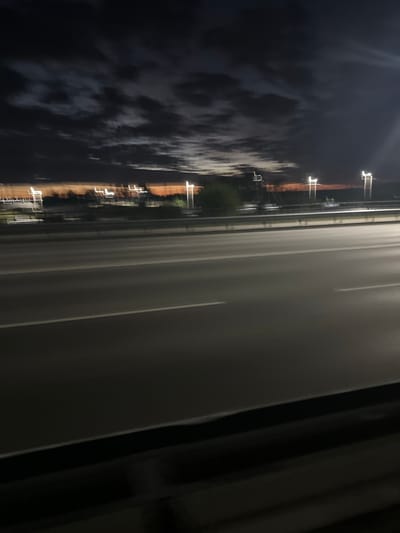 The image captures a motion-blurred view of a highway at night. The sky is dark with scattered clouds and a faint orange glow on the horizon, suggesting either sunset or distant city lights. Several tall lamp posts with bright lights are visible on the right side of the road, with more lights streaked across the distance on the left. The highway itself is paved, with visible lane markings. The foreground is dominated by a dark, blurred railing and the blurred road surface. There are no people or vehicles clearly discernible due to the motion blur. The location is identified as Заречный ауылдық округі, Kazakhstan.