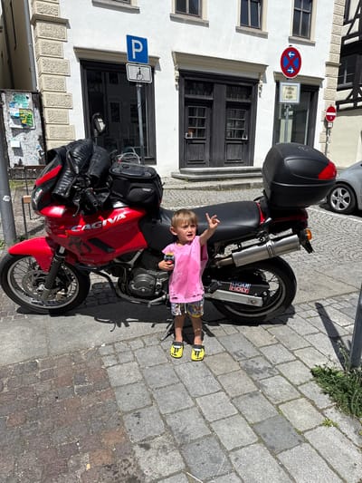 A daytime outdoor shot in Riedlingen, Germany features a child lying on a red Cagiva Ducati motorcycle parked on a cobblestone street. The child, wearing a pink shirt, patterned shorts, and yellow shoes, raises one hand in a "rock on" gesture. The motorcycle has black bags and a black trunk attached. In the background, a white building with dark-framed windows is visible. Road signs, including a parking sign and a no-parking sign, are mounted on the building. A portion of a gray car is also present in the upper background.