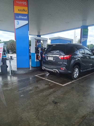 The image depicts a gray Isuzu MU-X SUV being refueled at a PTT Station in what appears to be Tha Ruea, Thailand. A gas station attendant wearing a dark blue uniform is operating the fuel pump on the passenger side of the vehicle. The fuel pumps are numbered, displaying prices for different types of fuel, including Gasohol 95, gasoline, and Diesel. The SUV's license plate number is กษ 9737. The background shows other fuel pumps, signage for Gasohol E20, Gasohol 91, Diesel, and a cashier booth labeled "Cashier". The ground is wet, suggesting recent rain.