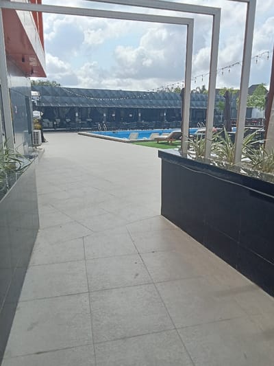 The image shows a view from a doorway looking out towards a swimming pool. The doorway is framed by a white metal structure. There is a black wall on the left side of the image and a black wall on the right side of the image. The pool is surrounded by a black tiled area. The pool is blue and there are some lounge chairs and tables in the surrounding area. The ground is tiled. There is a small patch of green grass visible to the left of the pool. The scene is an outdoor area located in the city of Ujevwu, Nigeria, at the coordinates latitude: 5.472, longitude: 5.817. There are no people visible in the image.