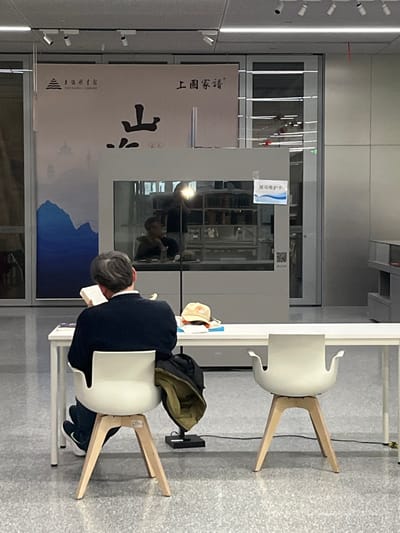 This image depicts an indoor scene within a modern library or exhibition space, specifically identified by a banner as the Shanghai Library. The location is stated to be the city of Yanqiao, China.
In the foreground, a man is seated with his back to the viewer at a long, white rectangular table. He has short, grey hair and is wearing a dark blue sweater or jacket over a lighter collared shirt, dark trousers, and white-soled sneakers with a visible Nike logo. He is actively reading an open book with white pages, held in both hands. A dark-colored jacket is draped over the back of his white, contemporary-style chair, which features light wooden, splayed legs. To his right, on the table, lies a light-colored baseball cap, possibly beige, with dark markings. A black electronic device or charger, connected by a cable, is on the floor beneath the table near his chair. Another identical white chair stands empty to the man's right at the same table.
The background features a large, reflective display case. The mirrored surface shows the reflection of another person, seated and appearing to look towards the left, along with a bright light source and a partial silhouette, possibly of the photographer. A white sign with blue text and border, reading "展项维护中" (Exhibit under maintenance), is affixed to the display case. Behind the display case, a large banner is displayed against a wall. It features Chinese characters "上海图书馆" (Shanghai Library) and "上圖家譜" (Shanghai Library Genealogy), along with a stylized landscape depicting blue and grey mountains, with the prominent character "山" (mountain) centrally displayed. The ceiling features multiple recessed spotlights. The floor throughout the space is a light grey, speckled material.
