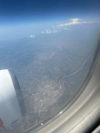 This is a photograph taken from an airplane window, looking down at a landscape. The image is oriented such that the horizon is vertical. In the foreground, a portion of the airplane's wing and fuselage is visible, with a reflective white surface. The middle ground shows a vast expanse of land, characterized by a patchwork of agricultural fields and a winding river. The fields are varied in color and shape, suggesting different crops or land use. A river snakes through the landscape, with its banks clearly defined. In the background, a clear blue sky dominates, transitioning to a hazy atmosphere closer to the horizon. Several wispy clouds are scattered across the sky and along the atmospheric haze. The lighting suggests it is daytime, likely with bright sunshine, as evidenced by the shadows and the clarity of the scene. There are no people or discernible text visible in the image. The view suggests a rural or semi-rural setting, possibly in Europe given the location context.