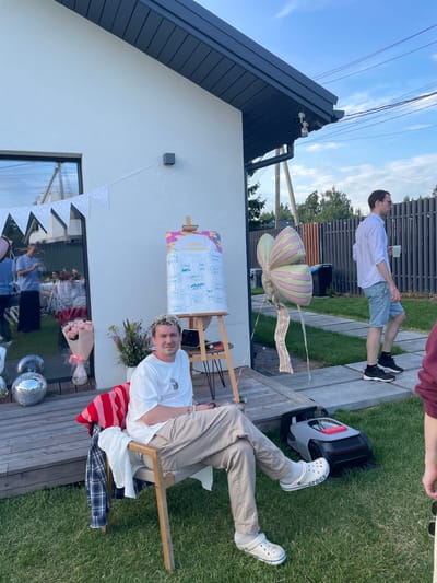 The image depicts an outdoor gathering at a modern white house in Simagino, Russia. The house features a dark grey roof and large windows. In the foreground, a man with curly blonde hair is seated in a chair, wearing a white t-shirt and beige pants. He is relaxed, with his legs crossed, and wearing white Crocs. A red and white striped blanket and a plaid shirt are draped over the chair. To the left, a bouquet of pink roses and disco balls are visible on a wooden deck. A whiteboard with handwritten notes and drawings, likely related to a birthday celebration, stands on an easel.

In the background, a man in a light blue shirt and shorts is walking on a paved path, holding a cup. A large, decorative balloon in the shape of a butterfly is tethered nearby. A robotic lawnmower is positioned on the grass. Several other people are seen in the background, some inside the house through the reflective windows and others standing on the grass. The scene is set against a backdrop of lush green grass, trees, and a fence, under a bright blue sky with some scattered clouds. The overall atmosphere appears festive and casual.