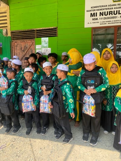 A group of Indonesian schoolchildren are gathered outdoors in front of a bright green wooden building. The children, primarily boys, are wearing green and yellow uniforms and white skullcaps, with some girls in yellow hijabs. Many of the children are carrying black backpacks and holding clear plastic bags filled with school supplies. The building appears to be part of an educational institution, with signage visible that includes "KEMENTERIAN AGAMA REPUBLIK INDONESIA" and "MI NURUL QOMARIYAH." The ground is covered in gravel. The overall scene suggests a school event or distribution of supplies.
