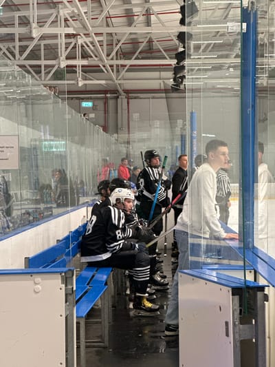 The image depicts an indoor ice rink, likely the Tnuvot Ice Arena in Tnuvot, Israel, viewed from behind a glass barrier. Several individuals, primarily young men wearing hockey gear, are seated on blue bleachers. Some are looking out onto the ice, while others appear to be conversing or resting. In the foreground, a young man in a white hoodie and blue jeans stands looking towards the ice, with his hand resting on the barrier. The environment shows a typical ice hockey facility with visible structural elements of the building above, lighting fixtures, and the wet ice surface reflecting the overhead lights. The overall scene suggests a break or pre-game period for the hockey players.