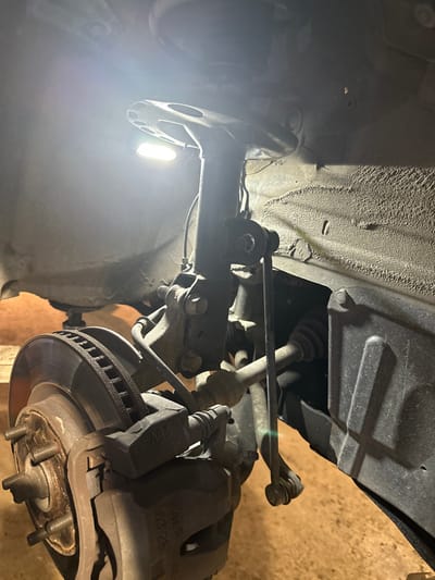 The image displays a detailed view of an automotive front suspension and braking system, likely undergoing maintenance or repair in Erma Reka, Bulgaria. The scene is illuminated by a bright, rectangular LED work light positioned in the upper left. Visible components include a heavily textured and somewhat soiled inner fender well, a multi-lug wheel hub, and a ventilated brake rotor exhibiting surface wear and minor corrosion. A brake caliper, bearing the embossed inscription "20V" and part numbers "ALR 63-28", is clamped onto the rotor. Directly above, a McPherson strut assembly is present, connecting to the vehicle's chassis structure, with a sway bar end link attached to the lower portion of the strut. A CV axle boot and shaft are partially visible towards the right, suggesting a front-wheel-drive configuration. The vehicle is elevated, with a jack stand partially visible supporting a lower component to the left. The ground surface, likely a garage floor, is visible at the bottom. No individuals or tools are visible in the frame.