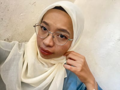 The media file is a vertically oriented, close-up, selfie-style photograph of a young person wearing a light-colored hijab and glasses. The subject's face is angled towards the camera, with their head tilted slightly to their left. They have dark eyes and are wearing subtle makeup, including lipstick. The person's right hand is visible near their face, gently touching their cheek or adjusting the hijab. They are wearing a blue garment on their left shoulder and a delicate silver bracelet on their wrist. The background appears to be a textured, light-colored surface, possibly a wall or floor. The lighting suggests an indoor setting, possibly during the day, given the even illumination. The overall mood is calm and contemplative. There is no visible text in the image.