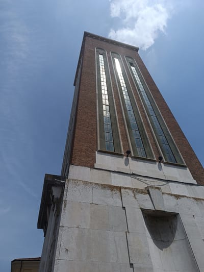 This image depicts a low-angle shot of the bell tower of the Church of Santa Maria Assunta in Casalmaggiore, Italy. The tower, constructed primarily of brick with stone detailing, features tall, narrow windows that reflect the bright sunlight and sky. The base of the tower is clad in large, light-colored stone blocks, some of which show signs of weathering and moss growth. A dark, recessed architectural feature is visible on the stone base. Electrical wires are strung across the facade of the stone base. The sky above is a clear blue with scattered wisps of white clouds. The overall impression is one of architectural grandeur against a bright, sunny day.