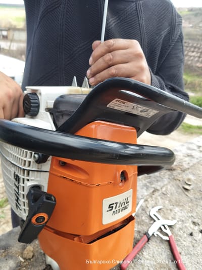 The scene is located in Balgarsko Slivovo, Bulgaria, captured on March 28, 2026, at 15:31. A Stihl MS 660 chainsaw, predominantly orange and white, is positioned on a light gray, textured concrete surface. A label on the chainsaw's top handle identifies it as "Andreas Stihl AG & Co. KG, D-71336 Waiblingen, MS 660, 2011". An individual's torso, clad in a dark, patterned sweater, is visible behind the chainsaw. The individual's right hand, with medium brown skin tone, manipulates a thin, metallic adjustment tool inserted into the chainsaw's upper mechanism. The left hand supports the chainsaw near its white housing. A black ELASTOSTART pull handle is visible on the chainsaw's left side. Adjacent to the chainsaw on the surface, a pair of slip-joint pliers with red handles rests. The background is out of focus, showing distant structures with reddish-brown roofs, sparse trees, and green, undulating terrain under an ambient light.