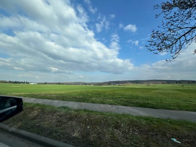 The image displays a panoramic view of an expansive rural landscape within the city of Stolberg, Germany, captured from a low perspective, likely from a moving vehicle. In the immediate foreground, the dark exterior of a vehicle is visible on the left, including a side mirror reflecting a blurred internal scene. A dark grey paved path or road runs horizontally across the lower portion of the frame, bordered by green and brown grassy verges.
The midground and background are dominated by vast, undulating green fields stretching towards a distant horizon. A subtle dirt track or path is observable traversing these fields into the distance. The horizon line reveals a mix of distant structures, including some light-colored buildings with industrial characteristics on the far left, interspersed with dense tree lines suggesting forested areas or hills.
The sky occupies the upper half of the image, featuring a dynamic composition of large, scattered white and grey cumulus clouds against patches of clear blue sky. In the upper right quadrant, bare tree branches with visible small buds extend into the frame. Several small, dark specks, consistent with birds, are observable in the mid-sky. A small piece of light-colored litter is present on the grassy verge in the bottom right corner. No individuals are visible.