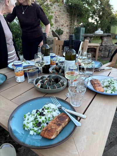 This image captures an outdoor meal scene in Crail, United Kingdom. A light brown, multi-panel wooden dining table is centrally placed, set with various food items and beverages, indicative of a social gathering.
Two women are partially visible on the left side of the frame. An older woman with grey hair is seen in profile, wearing a striped top and a dark cardigan. Adjacent to her, a younger woman with blonde hair, dressed in a dark brown knitted top and dark trousers, stands facing left, engaged in interaction.
The table holds multiple servings of food. In the foreground, a teal-blue ceramic plate with a brown rim contains a portion of cooked salmon fillet alongside white rice mixed with green peas and dark, possibly olive, pieces. A fork and knife with cream-colored handles are positioned on the plate. Another similar plate with salmon, rice, and peas is visible to the right. A grey ceramic bowl in the center of the table is filled with numerous maki rolls. Two green glass bottles of white wine, several wine glasses (some containing white wine, others empty), and clear drinking glasses are distributed across the table. Two colorful beverage cans are also present, one partially labeled "THE" and "LIME." A spice grinder or shaker with an orange top is visible near a can.
The environment suggests a garden or patio setting. A stone wall forms the background, interspersed with green foliage and trees. Wooden outdoor chairs are visible around the table. In the mid-ground background, a stone bench or table with a white decorative object and a green watering can are discernible. The lighting indicates natural outdoor light, likely late afternoon or early evening.