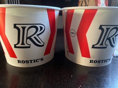 Two white paper buckets with red diagonal stripes and black lettering are placed side-by-side on a dark wooden table. The lettering on the left bucket says "ROSTIC'S". The lettering on the right bucket is partially obscured, but the word "ROSTIC'S" is also visible. A circular logo with "12+" inside it is also present on the right bucket. The buckets appear to be food containers, likely for fried chicken. The environment suggests an indoor setting, possibly a home or restaurant. The image is a close-up shot focusing on the buckets, with the background blurred. There is no information to indicate the location in the city of Akhtyrskiy, Russia.