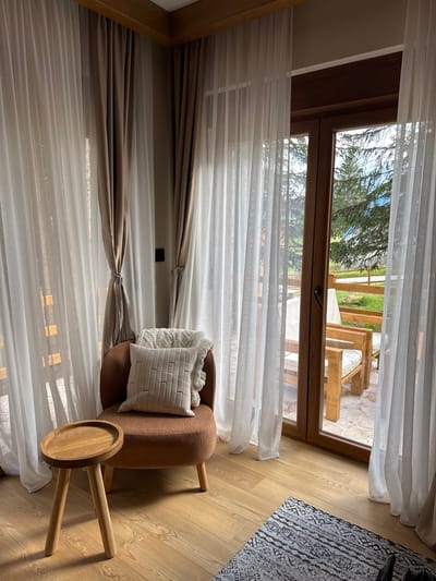 This is a medium-shot, eye-level photograph of a cozy corner in what appears to be a mountain lodge room in Kovačka Dolina, Montenegro. The room features light wood flooring and is decorated in warm, natural tones.
Dominating the left side of the frame is a plush, rust-colored armchair with a textured cream cushion and a knitted throw blanket draped over its back. To the left of the armchair sits a small, round, light-wood side table with three legs.
The room is framed by floor-to-ceiling curtains. Sheer white curtains are layered with heavier, taupe-colored curtains that are tied back with decorative fabric bands. These curtains partially obscure a large glass-paneled door and window that opens to an outdoor patio area.
Through the glass, we see two wooden lounge chairs with white cushions on a stone patio. Beyond the patio, there's a wooden fence, a gravel path, and lush green grass, all set against a backdrop of tall pine trees and a bright, slightly overcast sky. The overall impression is one of comfort and a connection to nature, likely a vacation rental or hotel room designed for relaxation. No people are visible in the scene.