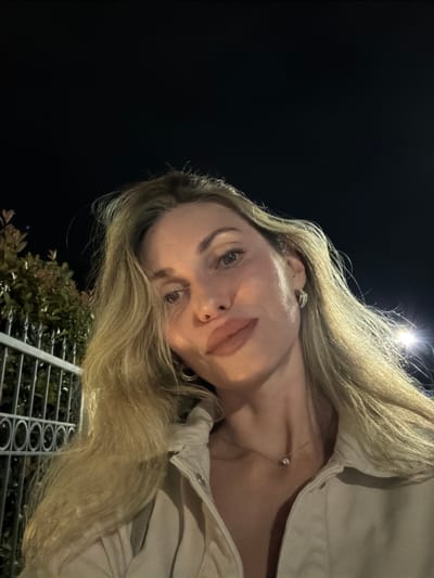 This is a tilted, low-angle, close-up selfie of a young woman at night in Adler, Russia. She has long, blonde hair that frames her face. She is wearing a light-colored jacket and a delicate necklace with a single pendant. Her eye makeup is subtle, and her lips are painted a nude shade. She is wearing hoop earrings.

The background is dark, suggesting it is nighttime. A bright light source, possibly a streetlamp, is visible in the upper portion of the image, casting a glow on her hair. In the lower part of the image, there is a glimpse of green foliage and a metal railing with a circular pattern. The woman's expression is neutral, and the lighting creates a soft, atmospheric mood. The overall impression is a casual, late-night portrait.