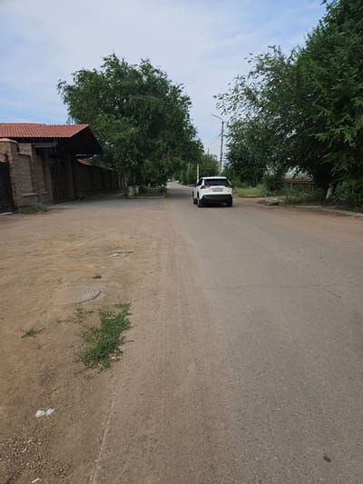 A white SUV with license plate "A042EX09" drives down an asphalt road lined with trees and some buildings. On the left side of the road, there is a fence, a building with a red tile roof, and a dirt area with a manhole cover. On the right side of the road, there are more trees and a wooden fence. The sky is cloudy. The image appears to be taken in Balqaş, Kazakhstan.