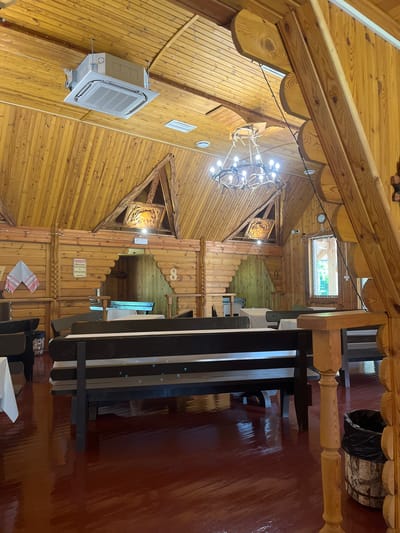 The image depicts the interior of a rustic wooden building, likely a restaurant or guesthouse, with a strong focus on natural wood elements and a cozy, traditional aesthetic. The walls and ceiling are constructed entirely of wood planks, showcasing their natural grain and knots. A large, elaborate chandelier made of antlers and bulbs hangs from the ceiling, providing warm lighting.
In the foreground, a dark wooden table with several chairs is visible. The floor is a dark red, highly polished wood that reflects the light. In the background, the building features multiple levels and architectural details, including arched wooden beams and decorative carvings on the walls. A small window with a view of greenery is positioned high up in one section of the ceiling.
An air conditioning unit is mounted on the wooden wall on the left side of the image. There are also some decorative items, such as a wooden carving on the wall and a woven mat hanging on a wooden partition. In one corner, a large wooden barrel lined with a black trash bag serves as a receptacle.
There is a sign on a wooden partition in the background with Cyrillic text, but it is partially obscured and difficult to read. The overall impression is one of a charming, rustic, and inviting space, possibly a rural establishment in Belarus. The lighting suggests it could be daytime, but the interior ambiance is warm and inviting. No people are visible in the image.