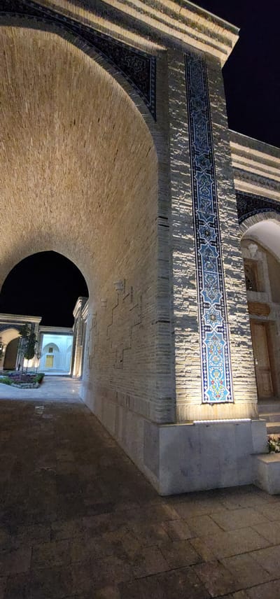 The image depicts an exterior view at night of an architectural complex with intricate Islamic tilework. The scene is illuminated by artificial lights, casting a warm glow on the textured brickwork and decorative tiles. In the foreground, a large arched structure made of light-colored brick dominates the left side of the frame. The archway opens into a dark, unlit space. To the right, a pillar is adorned with a vertical panel of blue and white mosaic tiles featuring floral and geometric patterns. The pillar is lit from below, highlighting its ornate decoration. Further into the background, a courtyard area is visible, leading to another building with a white facade and yellow doors, also illuminated. The ground is paved with stone or brick. The location is identified as being in the city of Satkak, Uzbekistan. There are no people visible in the image.