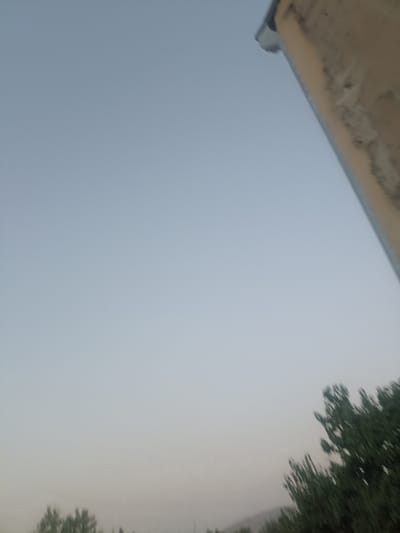 The image captures an outdoor scene with a pale blue sky dominating the frame. In the upper right corner, a portion of a light-colored, textured structure, possibly a building or a roof edge with a gutter, is visible. The lower portion of the image shows a blurred expanse of green foliage, suggesting trees or bushes. The overall lighting indicates it might be early morning or late evening. The scene appears to be in Aygezard, Armenia, given the context provided. No people or specific actions are discernible due to the blur and the composition of the image.