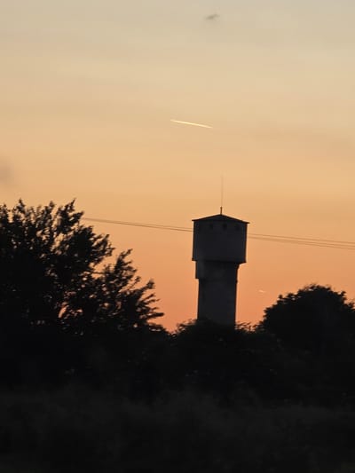 A landscape photograph in portrait orientation depicts a water tower silhouette in Karadjovo, Bulgaria, viewed against an orange-tinted sky at twilight. Darkened foliage frames the tower at the base and partially obscures its right side. Power lines run across the sky. An airplane contrail appears in the sky to the left of the tower.