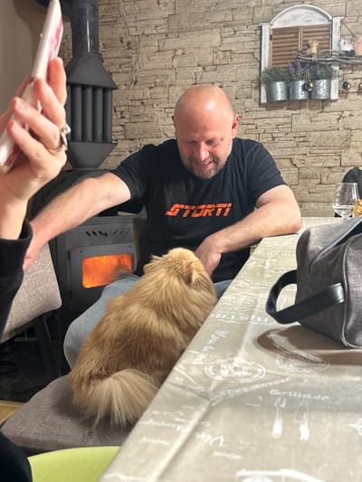 The image appears to be taken indoors, possibly in a private residence in Vinarovo, Bulgaria, though this is difficult to definitively confirm. A bald man wearing a black t-shirt with orange writing is seated at a table covered with a patterned tablecloth. Beside him, there is a small, reddish-brown dog. There is a small stove with a visible fire burning within it near the man and the dog. To the left, there is a decorative item on the wall and a black object that resembles a fireplace implement or decorative piece. In the foreground, a person's hands holding a mobile phone are visible. The scene suggests a cozy, domestic setting.