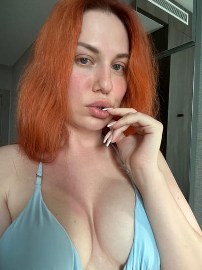 The image shows a close-up of a woman with bright orange hair. She is wearing a light blue halter top and has her hand near her mouth. Her fingernails are long and white. Her skin appears flushed, and there is a visible tan line on her chest and shoulder. The background is blurred but appears to be an indoor setting with a mirrored surface and a vent visible. The context of Avcılar, Turkey is not present in the image.