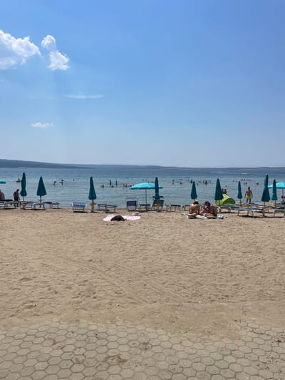 This is a ground-level, medium shot photograph of a crowded beach on a sunny day in Crikvenica, Croatia. The foreground is dominated by sand, with a hexagonal patterned walkway visible in the lower right corner. Several rows of blue lounge chairs and teal beach umbrellas are set up on the sand. In the mid-ground, the clear blue water of the Adriatic Sea is populated with numerous swimmers of various ages. Some individuals are wading, while others are swimming or playing in the water. A small boat is visible in the distance. Along the shoreline, several people are sunbathing on towels or chairs. Two women are seated on a towel in the center right of the frame, engaged in conversation. The background consists of a hazy, distant coastline and a clear blue sky with a few wispy clouds. The overall atmosphere is one of leisure and recreation.