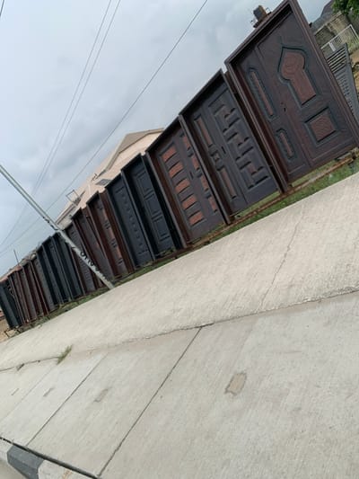 The image presents an outdoor display of approximately 18 to 20 large, dark-colored doors, likely made of wood or metal, arranged in a linear fashion along a strip of ground in Igando Oloja, Nigeria. The doors exhibit various intricate designs, including geometric patterns, recessed panels, and detailed carvings, with one door on the far right featuring an arched motif and script. They appear to be freestanding or supported by unseen structures. In the foreground, a wide, light gray concrete pavement composed of segmented slabs extends across the lower half of the frame, showing minor cracks and sparse patches of green grass. A black and white striped curb is visible at the bottom left. Overhead, a utility pole on the left supports multiple dark wires against a heavily overcast, gray sky. Behind the doors, a light-colored building with a pitched roof is partially visible, and a dark water storage tank sits atop a distant structure on the right horizon. No individuals are present, and no actions are observed.