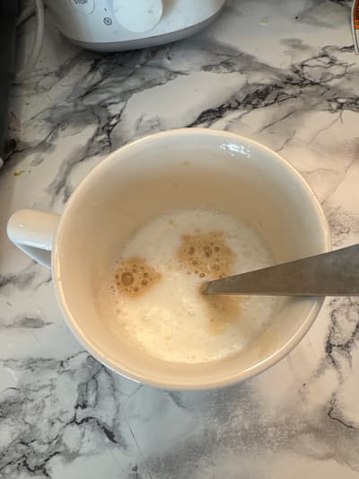 This is a top-down, close-up shot of a white ceramic mug filled with a frothy, light brown beverage, likely coffee or a similar drink. A silver spoon is partially submerged in the liquid, indicating that the drink is being stirred or has just been prepared. The foam on the surface of the drink has small bubbles and some darker, concentrated areas, suggesting the presence of espresso or coffee.

The mug is placed on a countertop with a marble-like pattern, featuring veins of black and grey against a white background. To the left of the mug, a portion of a white electronic device is visible. It has a circular dial and buttons, one labeled "STOP" and another with a timer icon. This device appears to be a kitchen appliance.

The lighting suggests it could be daytime, possibly indoors in a kitchen or dining area. There are no explicit indicators of the weather or specific time of day beyond general daylight. The scene is clean and tidy, with no visible mess. The overall mood is calm and domestic, possibly capturing a moment of preparation for a beverage.

There is some visible text on a sticker partially visible at the top left of the image, but it is mostly obscured and illegible. The text on the appliance buttons is clear: "STOP" and a circular symbol that appears to be a timer or clock.