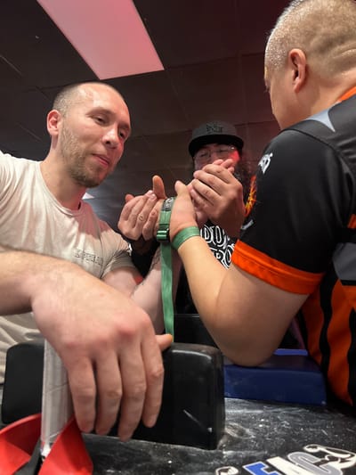 Three men are arm wrestling. One man, wearing a white shirt, is seated at the table with his arm secured by a green strap. Another man in a black and orange shirt is next to him. A third man, wearing a baseball cap and glasses, is watching them. The arm wrestling table is black and white.
