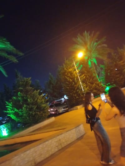 The image depicts two women at night. One woman, in the foreground and on the right, is holding a mobile phone and appears to be taking a picture or recording a video. She has dark, long hair and is wearing a light-colored top and dark bottoms. The other woman, on the left, is facing away from the camera and also holding a mobile phone. She has dark hair and is wearing a dark top and blue jeans.

The setting appears to be an outdoor area at night, possibly a park or a public space in Rudəkənar, Azerbaijan. There are palm trees and other dense green foliage, some illuminated with green lights. A street lamp with a bright yellow light is visible, as well as power lines. In the background, there is a silver car parked on a street. The ground is paved with a yellowish-orange surface, and there are raised concrete or stone structures, possibly planters or decorative elements. The overall lighting suggests it is after dark, with artificial lights illuminating the scene.