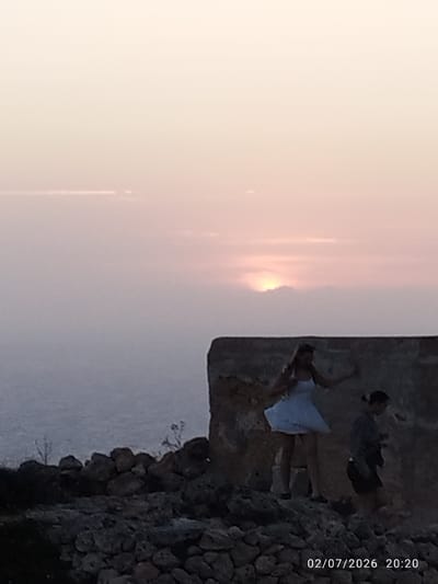 Two individuals are positioned on a rocky outcrop overlooking a body of water at sunset. A person in a white dress stands on the left, with their arm raised. To the right, another person in dark clothing walks away from the camera. The background features a hazy sunset with the sun partially obscured by clouds. The foreground consists of stacked rocks, forming a rough wall. The image is dated "02/07/2026 20:20".