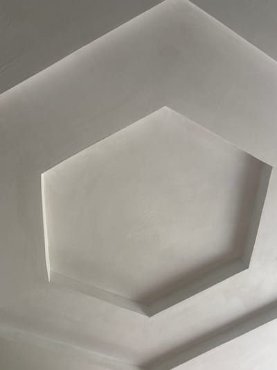 The image shows a close-up view of a white, textured ceiling with a hexagonal recessed design. The lighting creates shadows and highlights that emphasize the geometric pattern and the texture of the material. There are no people, objects, or discernible activities taking place. The scene is abstract and architectural, focusing solely on the form and texture of the ceiling. The time of day and weather are not discernible from the image, nor are there any specific location cues within the frame, other than the provided context of Tutaev, Russia. The overall mood is neutral and calm, due to the monochromatic color scheme and the simple geometric design. There is no visible text in the image.
