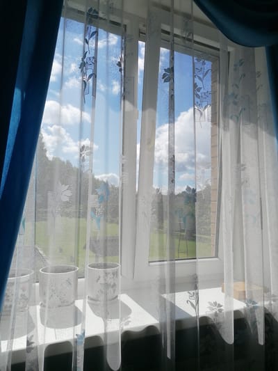 The image depicts a window with sheer white curtains patterned with blue flowers and geometric shapes, overlaid with a partially drawn blue curtain on the left. Through the window, a bright sky with scattered clouds is visible, along with a green landscape of trees and grass. On the windowsill, two white cylindrical planters with floral designs are present. The scene appears to be an interior view looking outwards, likely from a residential building in Stropi, Latvia, given the context provided. The lighting suggests it is daytime.